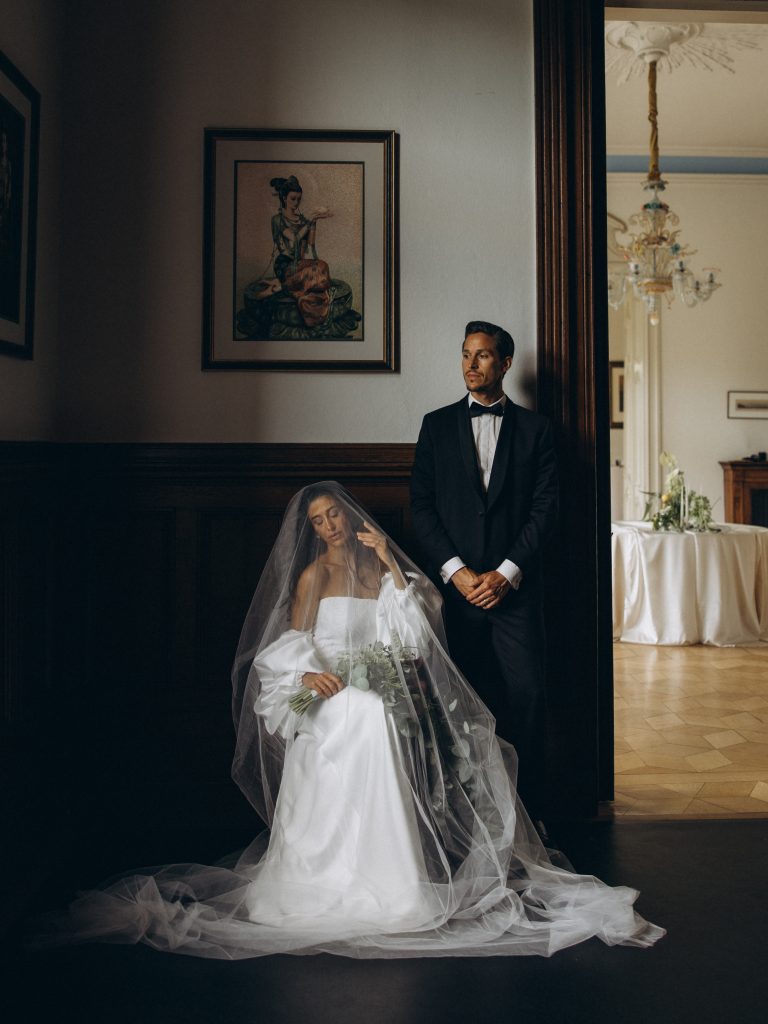 Braut sitzt unter ihrem Schleier und der Bräutigam steht neben ihr.